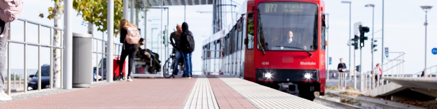 Helle Außenaufnahme eines Bahnsteigs in Düsseldorf: Eine rote U-Bahn der Linie U77 fährt an einer Haltestelle mit wartenden Fahrgästen ein. Im Vordergrund sind weiße taktile Leitstreifen auf dem Pflaster erkennbar; im Hintergrund die Pylone der Oberkasseler Brücke unter hellem Himmel.