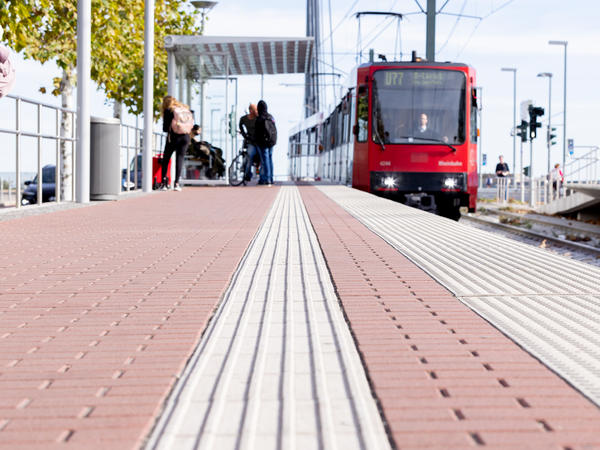 Blick aus tiefer Perspektive über einen Bahnsteig in Düsseldorf: Weiße taktile Leitstreifen im rötlichen Pflaster führen zentral ins Bild. Im Hintergrund wartende Fahrgäste und eine einfahrende rote U-Bahn der Linie U77 vor der Kulisse der Oberkasseler Brücke.