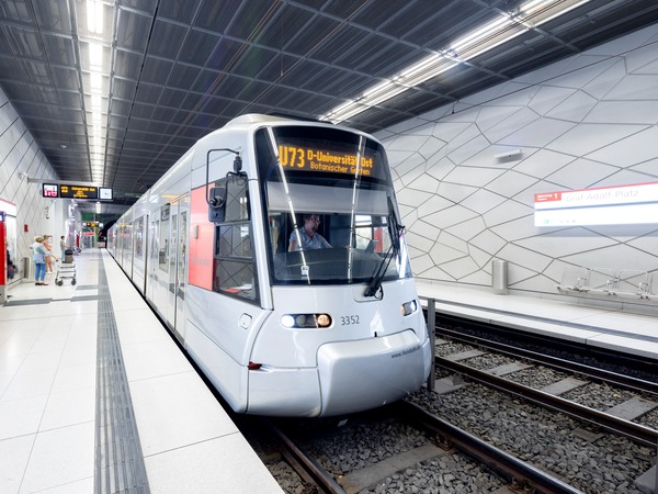 Das Bild zeigt eine stilisierte Illustration in kräftigem Rheinbahn-Rot. Im Vordergrund stehen vier diverse Menschen – zwei Frauen und zwei Männer – die miteinander im Gespräch sind. Hinter ihnen fährt eine moderne, rot-weiße Niederflurstraßenbahn mit dem Ziel „Düsseldorf“. Im Hintergrund sind bekannte Wahrzeichen der Stadt zu sehen, darunter der Rheinturm und die Lambertuskirche. Die Menschen wirken interessiert und zugewandt, so dass der Eindruck entsteht, sie tauschen sich über die Bahn oder das Thema Mobilität in Düsseldorf aus.